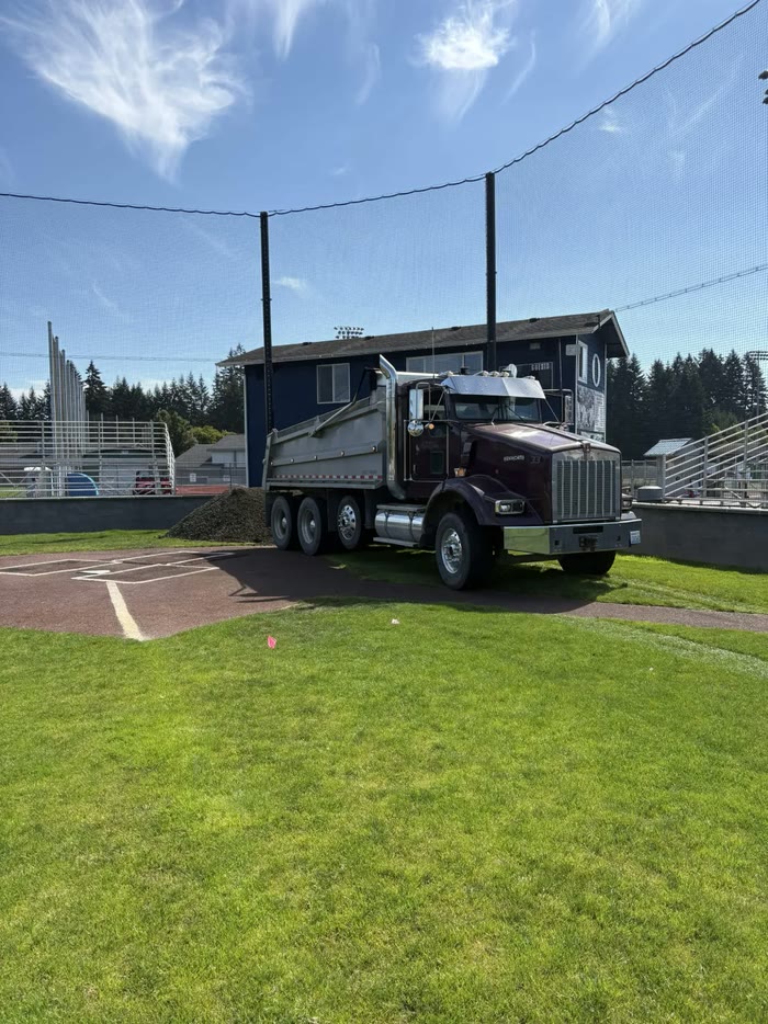 A maroon Kenworth dump truck parked on the grass beside home plate at a ball field, a pile of fill next to it.