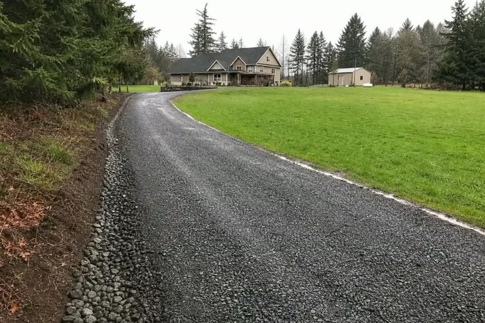 A long curving driveway of fresh dark crushed rock edged with river stones, leading past a green field to a farmhouse.