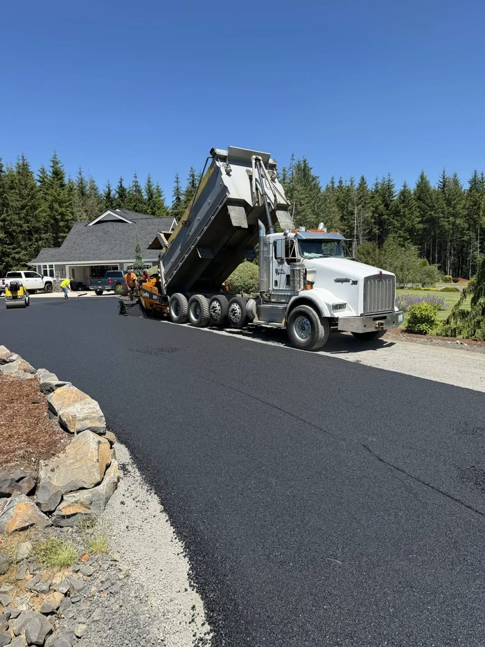 A Barnes Hauling dump truck tips asphalt into a paving machine on a new driveway, a house and fir trees behind.