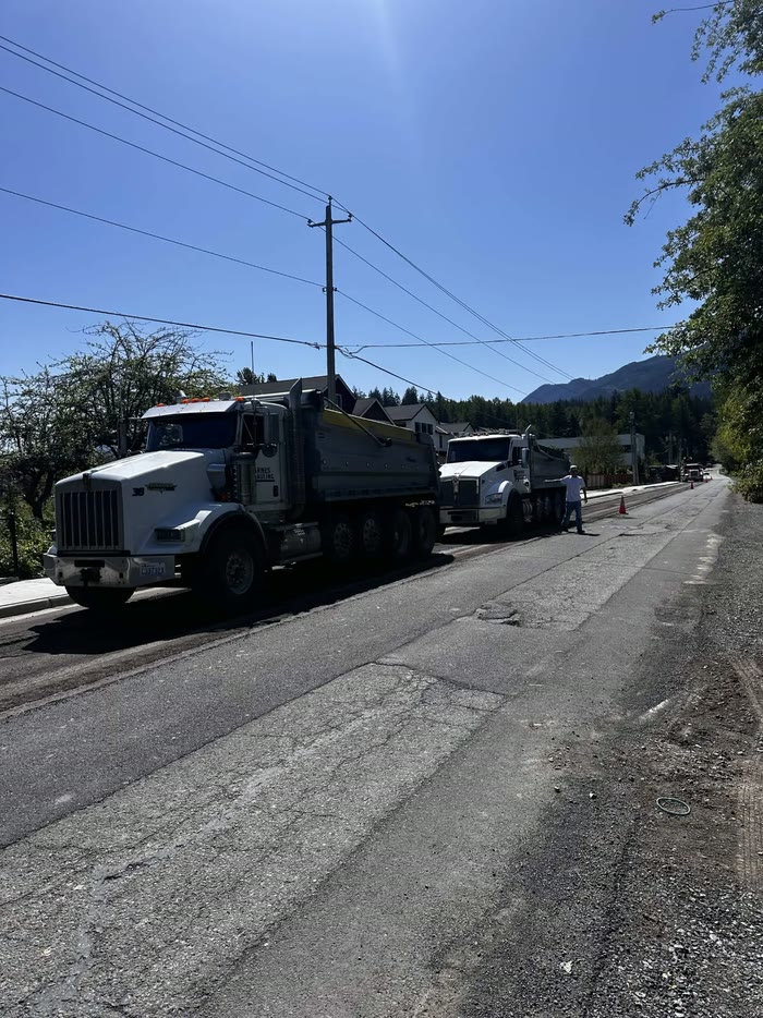 Two Barnes Hauling dump trucks parked along a road being resurfaced, a worker waving the next truck in.